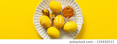 Bright yellow Easter eggs arranged on a white plate with quail eggs and a small bowl of turmeric add a vibrant touch to the holiday decorations and festivities, a way to dye eggs yellow, banner 134930531