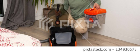 A person is getting ready to vacuum a room. They are attaching a container to the vacuum cleaner while kneeling on the floor. A plant is visible in the background, banner A person is getting ready to vacuum a room. They are attaching a container to the vacuum cleaner while kneeling on the floor. A plant is visible in the background, banner 134930575