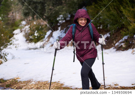 Woman Hiking in Snowy Trail with Trekking Poles, Maroon Jacket, and Backpack 134930694