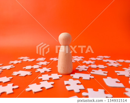 Wooden peg doll surrounded by white jigsaw puzzle pieces on an orange background. Wooden peg doll surrounded by white jigsaw puzzle pieces on an orange background. 134931514