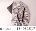 Magnifying glass and a pen resting on a fan of US one-hundred-dollar bills. 134931517