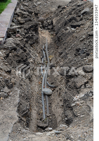 A section of dug trenches at edge of the carriageway of an asphalt road, under which black plastic pipes were laid for water supply or drainage. Repair and restoration works of underground utilities A section of dug trenches at edge of the carriageway of an asphalt road, under which black plastic pipes were laid for water supply or drainage. Repair and restoration works of underground utilities 134933947