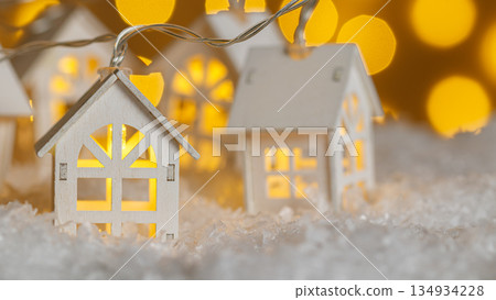 Christmas wooden houses garlands with lights in the snow on glowing lights bokeh background Christmas wooden houses garlands with lights in the snow on glowing lights bokeh background 134934228