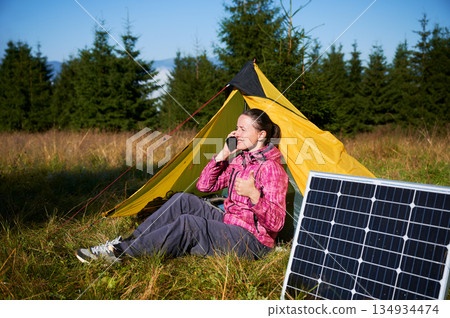Happy woman sits in tent, talking on her phone and giving thumbs up. Solar panel, highlighting use of renewable energy and eco-friendly camping. On background lush green trees and clear blue sky. Happy woman sits in tent, talking on her phone and giving thumbs up. Solar panel, highlighting use of renewable energy and eco-friendly camping. On background lush green trees and clear blue sky. 134934474