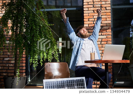 Man stretches with arms overhead while working on laptop at outdoor round table. Solar panel nearby, emphasizing sustainable, modern eco-friendly workspace that combines technology and sustainability. Man stretches with arms overhead while working on laptop at outdoor round table. Solar panel nearby, emphasizing sustainable, modern eco-friendly workspace that combines technology and sustainability. 134934479