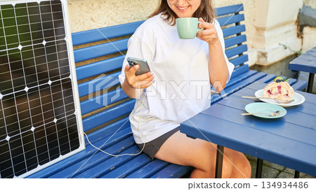 Girl drinking coffee while phone charging from solar panel. Cropped view of happy woman sitting next to portable solar battery charger. Integration of sustainable renewable energy into everyday life. Girl drinking coffee while phone charging from solar panel. Cropped view of happy woman sitting next to portable solar battery charger. Integration of sustainable renewable energy into everyday life. 134934486