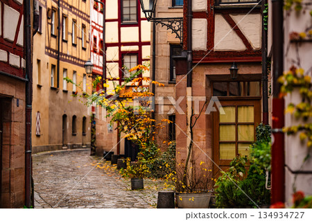 Charming Old Street In Nuremberg: Narrow Cobblestone Alley With Historic Half-Timbered Houses And Autumn Leaves Creating A Warm Medieval Atmosphere Charming Old Street In Nuremberg: Narrow Cobblestone Alley With Historic Half-Timbered Houses And Autumn Leaves Creating A Warm Medieval Atmosphere 134934727