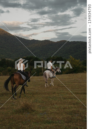 Two riders on horses explore a grassy field near mountains during late afternoon Two riders on horses explore a grassy field near mountains during late afternoon 134934793