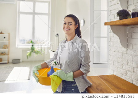Woman housewife in gloves with cleaning tools and rags, standing in kitchen and looking at camera. Woman housewife in gloves with cleaning tools and rags, standing in kitchen and looking at camera. 134935585