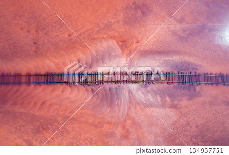 Aerial of a railroad tracks crossing a pink-hued lake. The perspective offers a unique view of the railway lines submerged in the shallow waters of the lake. Burlinskoe Lake, located in Bursol. Aerial of a railroad tracks crossing a pink-hued lake. The perspective offers a unique view of the railway lines submerged in the shallow waters of the lake. Burlinskoe Lake, located in Bursol. 134937751