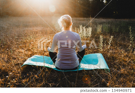 Woman practicing yoga and meditation on nature sitting in lotus pose on yoga mat, relaxed with closed eyes. Mindful meditation concept. Wellbeing. Sunset meditation. Woman practicing yoga and meditation on nature sitting in lotus pose on yoga mat, relaxed with closed eyes. Mindful meditation concept. Wellbeing. Sunset meditation. 134940971