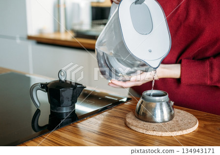 Person pours filtered water from pitcher into base of moka pot on wooden counter. Manual espresso routine, elevated home brewing, artisanal coffee gear, precision coffee making. Person pours filtered water from pitcher into base of moka pot on wooden counter. Manual espresso routine, elevated home brewing, artisanal coffee gear, precision coffee making. 134943171