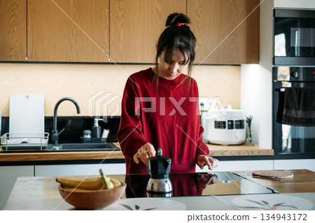 Woman operates induction stove to brew coffee with moka pot in modern kitchen. Induction stove brewing, modern kitchen appliances, smart home living. 134943172