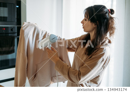 Woman uses portable garment steamer on sweatshirt in kitchen. Lightweight travel gear, business trip essentials, garment care. Woman uses portable garment steamer on sweatshirt in kitchen. Lightweight travel gear, business trip essentials, garment care. 134943174