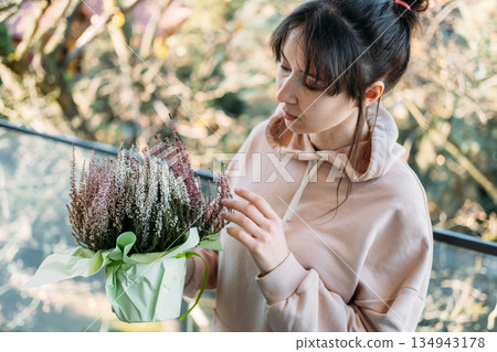 Woman examines potted heather plant on sunlit balcony. Biophilia, indoor outdoor living, balcony plants, heather flower. 134943178
