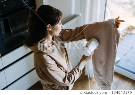Woman uses handheld garment steamer on trousers in kitchen. Smart home appliances, time-saving technology, modern laundry care. Woman uses handheld garment steamer on trousers in kitchen. Smart home appliances, time-saving technology, modern laundry care. 134943179