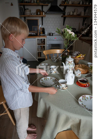 Young boy sets a tea table at home, placing cups and spoons on a tablecloth in a cozy kitchen with soft natural light 134944579