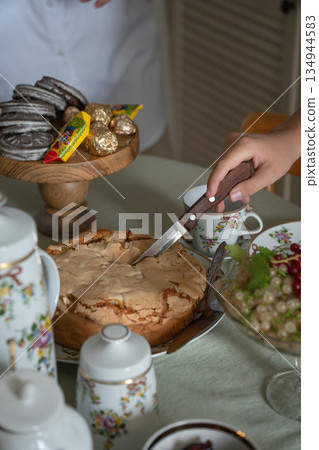 Hands cut a homemade apple charlotte on a tea table with floral teapot, cups, cookies, and bowls of red and white currants in soft natural light 134944583