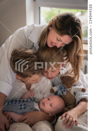 A mother embraces her three young sons, smiling warmly by the window as they sit together on the bed A mother embraces her three young sons, smiling warmly by the window as they sit together on the bed 134944588