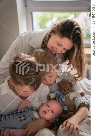 A mother embraces her three young sons, smiling warmly by the window as they sit together on the bed 134944589