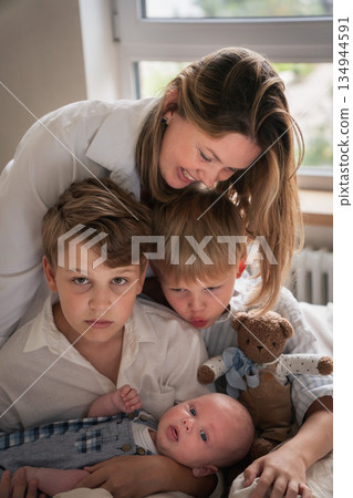 A mother embraces her three young sons, smiling warmly by the window as they sit together on the bed A mother embraces her three young sons, smiling warmly by the window as they sit together on the bed 134944591