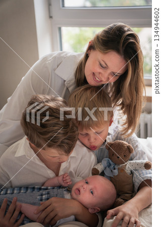 A mother embraces her three young sons, smiling warmly by the window as they sit together on the bed A mother embraces her three young sons, smiling warmly by the window as they sit together on the bed 134944602