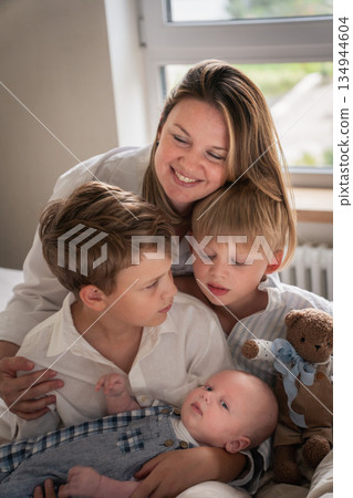 A mother embraces her three young sons, smiling warmly by the window as they sit together on the bed A mother embraces her three young sons, smiling warmly by the window as they sit together on the bed 134944604