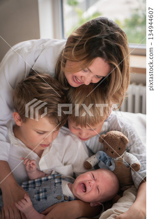 A mother embraces her three young sons, smiling warmly by the window as they sit together on the bed A mother embraces her three young sons, smiling warmly by the window as they sit together on the bed 134944607