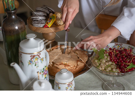 Hands cut a homemade apple charlotte on a tea table with floral teapot, cups, cookies, and bowls of red and white currants in soft natural light Hands cut a homemade apple charlotte on a tea table with floral teapot, cups, cookies, and bowls of red and white currants in soft natural light 134944608