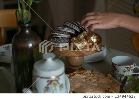 Hand reaches for sweets on a wooden stand beside homemade apple charlotte, gingerbread cookies, candies, and a floral tea set on a cozy table 134944622