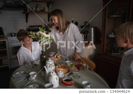 Mother laughs with her children around kitchen table set with homemade cake, tea cups, flowers, desserts, capturing lively family moment Mother laughs with her children around kitchen table set with homemade cake, tea cups, flowers, desserts, capturing lively family moment 134944624