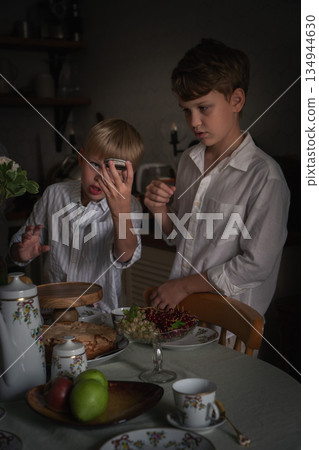 Two young brothers stand by home tea table with apple charlotte, berries, fruit, floral tea set, sharing candid moment in soft natural kitchen light Two young brothers stand by home tea table with apple charlotte, berries, fruit, floral tea set, sharing candid moment in soft natural kitchen light 134944630