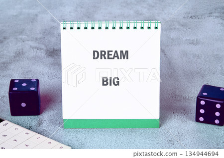 A spiral notebook rests on a textured surface displaying the words DREAM BIG. Two dice and a calculator are also placed near the notebook 134944694
