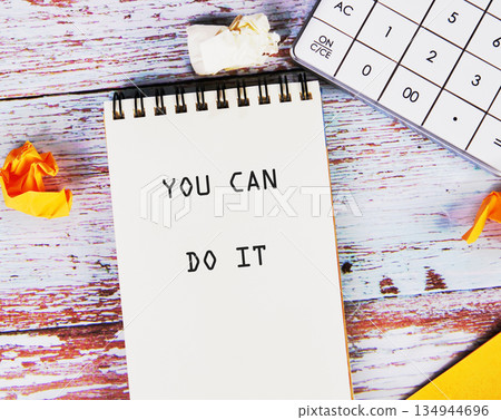 A notebook displays the message YOU CAN DO IT near a calculator and crumpled paper on a rustic wooden desk. Encouragement is in the air 134944696
