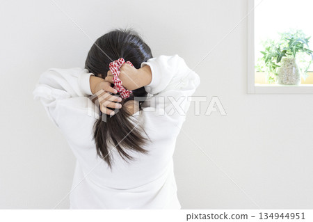 A girl (5 years old, Japanese, black hair) trying to tie her hair in a ponytail. 134944951