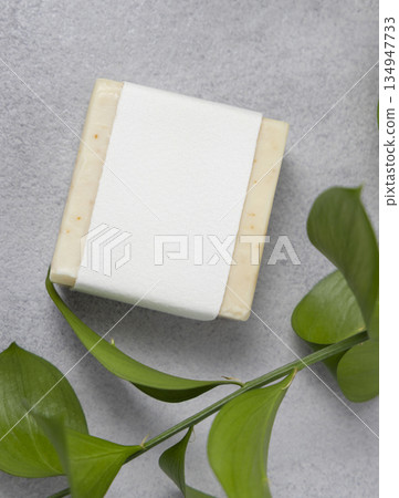 Soap with blank label on white bath towel near green leaves on grey stone top view, mockup 134947733