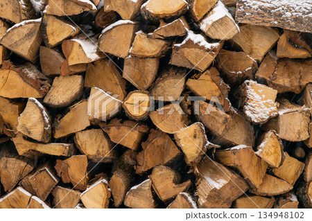 A close-up texture of dry chopped wood logs stacked outdoors during winter. Light snowflakes cover the surface of the timber. A close-up texture of dry chopped wood logs stacked outdoors during winter. Light snowflakes cover the surface of the timber. 134948102