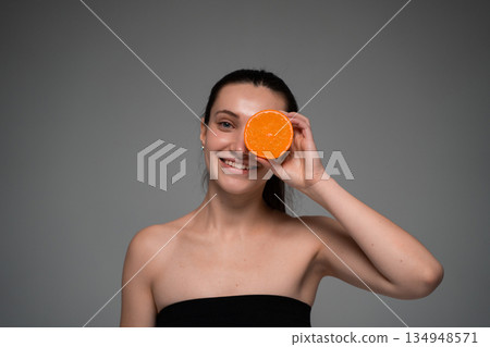 Young woman holding fresh orange slices over eyes in minimalist studio portrait, creative citrus concept for skincare, wellness, beauty and healthy lifestyle 134948571