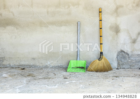 A traditional grass broom and a green plastic dustpan filled with debris stand against a weathered A traditional grass broom and a green plastic dustpan filled with debris stand against a weathered 134948828