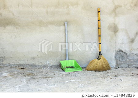 A traditional grass broom with a golden handle and a modern green plastic dustpan, holding debris, lean against a textured A traditional grass broom with a golden handle and a modern green plastic dustpan, holding debris, lean against a textured 134948829