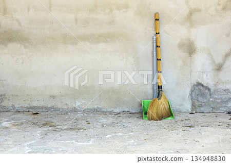 A traditional grass broom with a black-wrapped handle and a bright green plastic dustpan lean against a textured A traditional grass broom with a black-wrapped handle and a bright green plastic dustpan lean against a textured 134948830