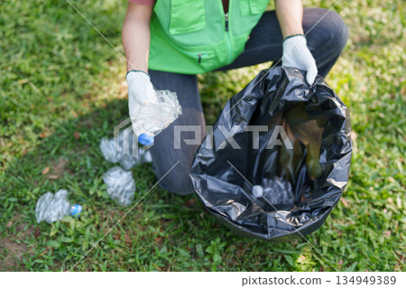 Adult volunteer collects plastic bottles wearing protective gloves clean up plastic waste on green grass outdoor park environmental protection 134949389