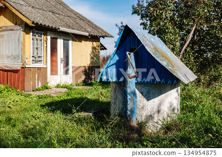 Old abandoned water well with rusty metal roof and wooden house in the background Old abandoned water well with rusty metal roof and wooden house in the background 134954875