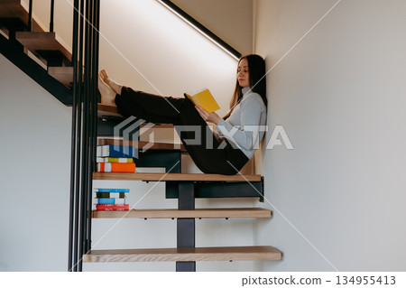 Young woman reading paper book in yellow cover sitting on modern wooden stairs in cozy minimalist home interior Calm weekend mood. Clever girl indoor. Young woman reading paper book in yellow cover sitting on modern wooden stairs in cozy minimalist home interior Calm weekend mood. Clever girl indoor. 134955413