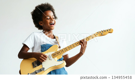 Young girl with curly hair joyfully playing electric guitar, wearing casual clothing, in a bright studio setting, showcasing musical talent and passion for music 134955458