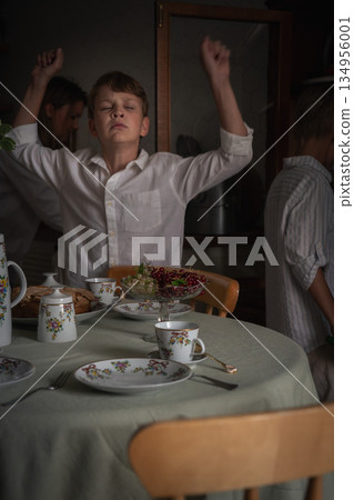 Playful boy raises his arms and makes a funny face at a home tea table with floral teaware, berries, and homemade apple cake in soft kitchen light Playful boy raises his arms and makes a funny face at a home tea table with floral teaware, berries, and homemade apple cake in soft kitchen light 134956001