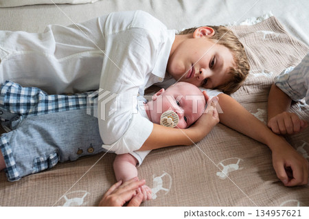Two brothers lie on a bed hugging a newborn baby with a pacifier, soft daylight, neutral colors, calm and tender sibling bonding at home 134957621