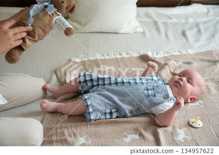 Newborn baby lies on soft blanket on bed while a hand holds a plush teddy bear nearby, neutral colors, gentle daylight, calm and cozy home atmosphere Newborn baby lies on soft blanket on bed while a hand holds a plush teddy bear nearby, neutral colors, gentle daylight, calm and cozy home atmosphere 134957622