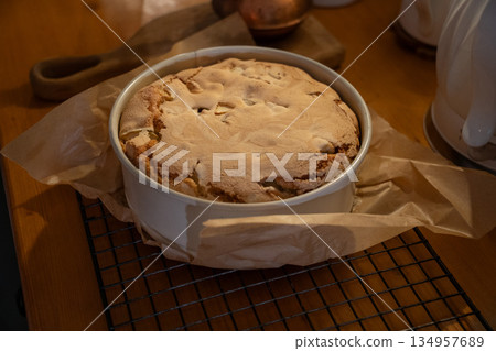 Freshly baked apple pie with golden crust cools in a baking dish on parchment paper, warm tones, cozy homemade baking atmosphere 134957689