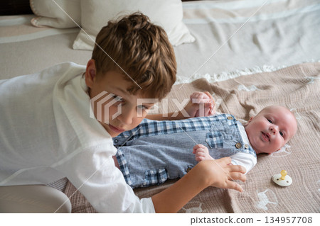 Older brother gently touches a newborn baby lying on a bed at home, soft daylight, neutral colors, tender sibling interaction 134957708